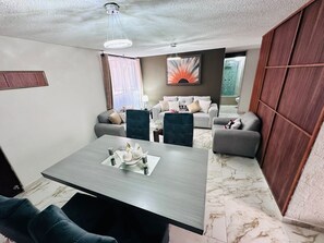 Ein modernes Wohnzimmer mit Essbereich, grauen Sofas, einem dunkel gepolsterten Sessel und einem großen abstrakten Gemälde an der Wand.