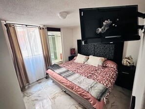 Ein Schlafzimmer mit Bett, Kopfstütze, Nachttisch, Fenster mit Vorhängen und Blick auf einen Balkon.
