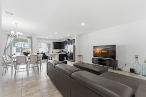 Ein modernes Wohnzimmer mit einer großen Eckcouch, einem Essbereich mit Tisch und Stühlen, einer Küche mit dunklen Schränken und einem flachbildfernseher an der Wand.
