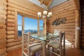 L’intérieur d’une cabane en rondins, avec une table à manger en verre, des chaises en bois, un lustre et une vue par les fenêtres.