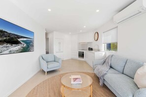 Ein modernes Wohnzimmer mit Sofa, Sessel und Couchtisch, einem Küchenbereich und einem großen Fenster mit Meerblick.