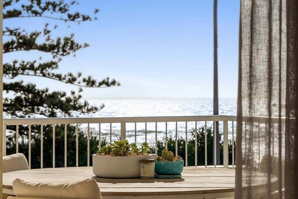 Un balcón con vistas al mar, una mesa con cojines y plantas en macetas.