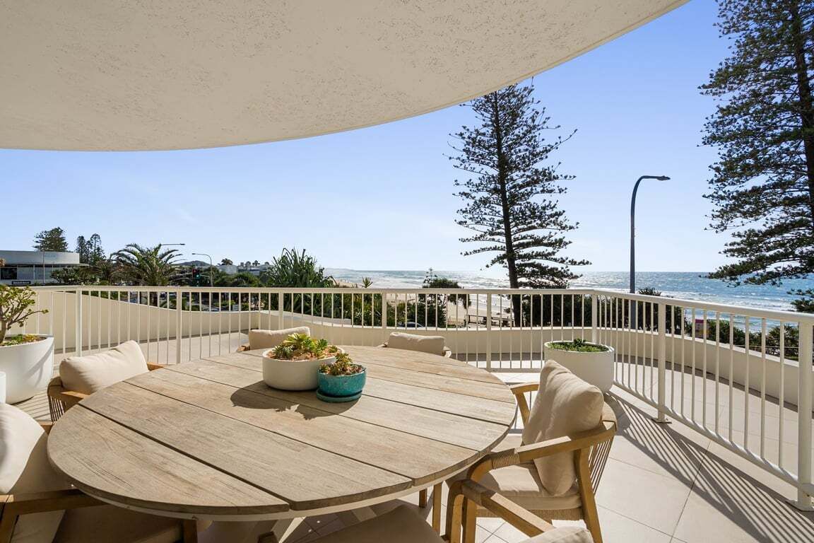 Et træbord med stole og potteplanter på en balkon med udsigt over strand og hav.