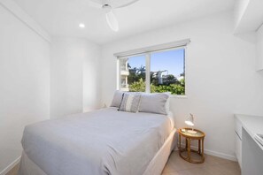 Ein Schlafzimmer mit Bett, Nachttisch mit Lampe, Fenster mit Blick ins Grüne und Deckenventilator.
