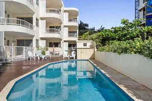 Ein Swimmingpool mit weißen Liegestühlen, einer Holzterrasse und einem Gebäude mit Balkonen.
