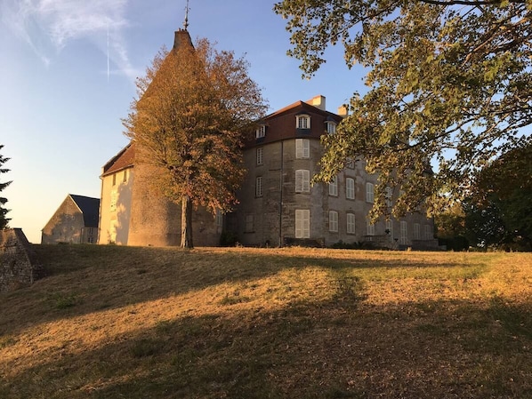 Un grand bâtiment en pierre, doté de nombreuses fenêtres et d’une tour imposante, entouré d’arbres et d’une zone herbeuse.