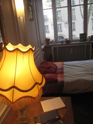 A room with a bed, a lamp, a window with a view of buildings, and a radiator. - Sofa bed near Parc de la tête d'or (Lyon)