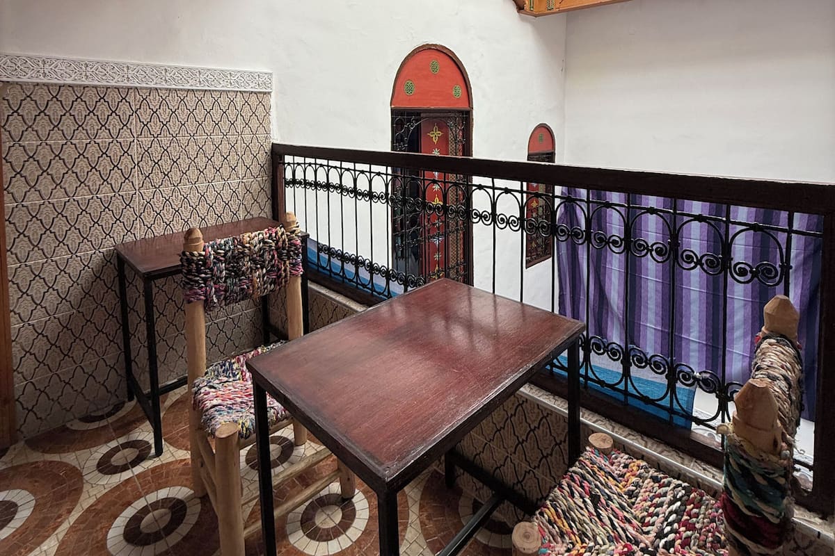 A room with a wooden ceiling, a patterned floor, a small table, a bench, and a view through a window with a metal railing.
