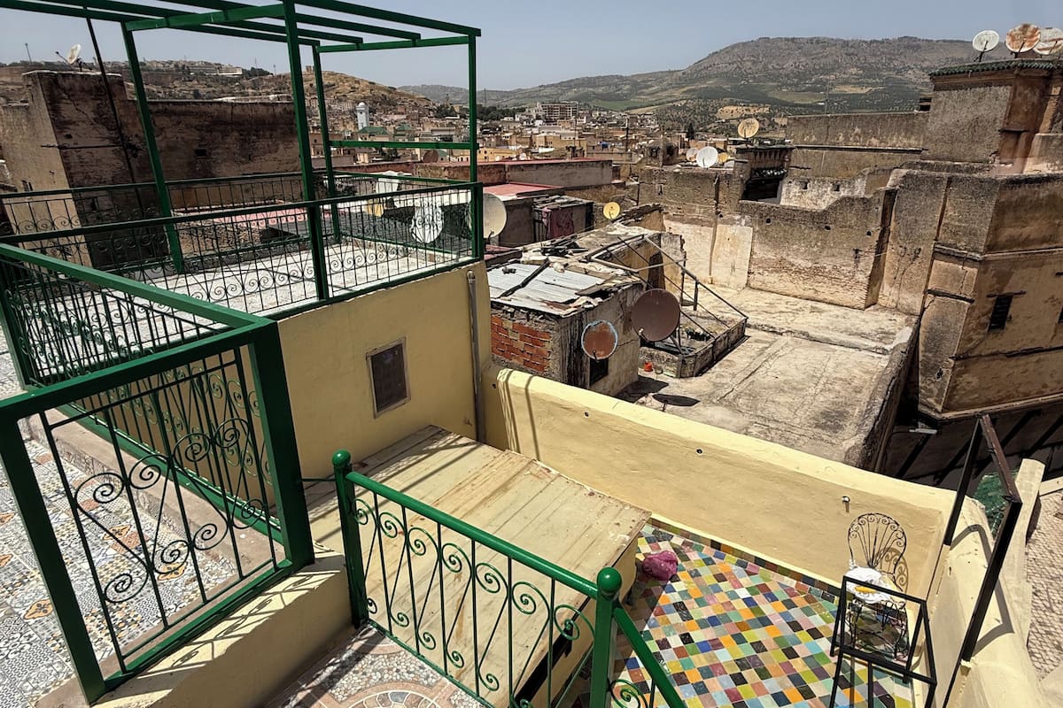 A rooftop terrace with a view of a cityscape, a tiled floor, and a green metal railing.