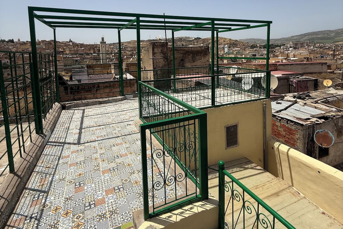 A rooftop terrace with a tiled floor, green metal railings, and a view of a cityscape.