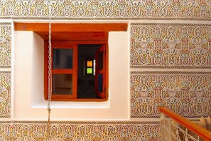 A room with a window featuring intricate wall patterns, a wooden door, and a chain hanging from the ceiling. - Riad AL HANA (Fes)