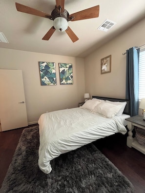 Une chambre à coucher avec un lit, un ventilateur de plafond, deux œuvres d’art murales, une porte et une fenêtre avec des rideaux.
