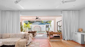 A spacious living area with a sofa, coffee table, and dining set. A ceiling fan is visible, and the room opens to a pool area. - The Beachside House with Pool & Walk to Beach (Burleigh Heads)