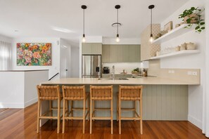 A modern kitchen with a central island, bar stools, pendant lighting, and a stainless steel refrigerator. - The Beachside House with Pool & Walk to Beach (Burleigh Heads)