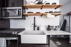 Una cocina moderna con microondas integrado, estufa y fregadero. Hay repisas de madera sobre el fregadero con platos y una tetera.