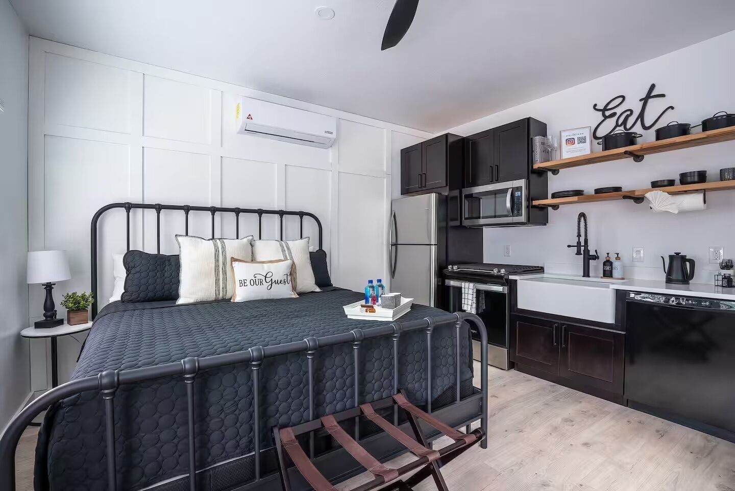 Ein gemütliches Schlafzimmer mit Bett, Nachttisch, Lampe und einem Wandgerät zur Klimatisierung. Angrenzend befindet sich eine Küchenzeile mit Mikrowelle, Herd und Regalen.