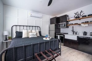 Un dormitorio pequeño con cama, mesita de noche, lámpara y aire acondicionado instalado en la pared. Junto a él, una kitchenette con microondas, estufa y estanterías.