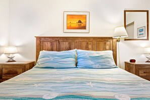 Una cama con colcha azul y blanca estampada, cabecera de madera, dos lámparas de noche, un cuadro enmarcado en la pared y una mesita de noche con un reloj.