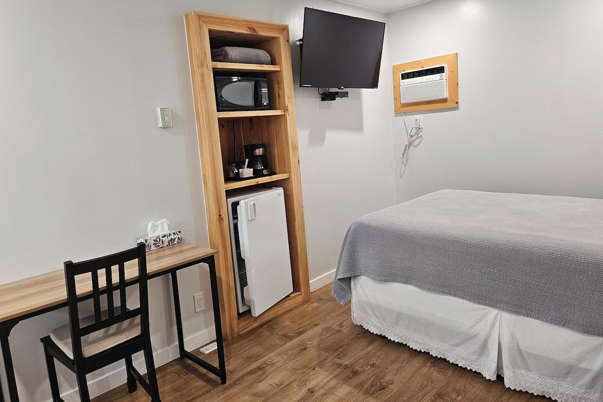 Quarto com cama, escrivaninha com cadeira, micro-ondas, frigorífico, televisão e ar condicionado de parede.