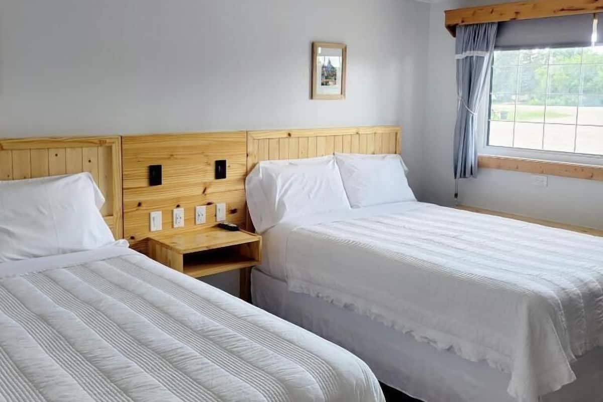 Quarto com duas camas, cabeceiras de madeira, mesa de cabeceira, quadro pendurado na parede e janela com cortinas.