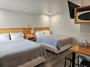 A hotel room with two beds, a wooden headboard, a small table, a TV mounted on the wall, and a brick-patterned wall. - Quest Motel (Whitewood)