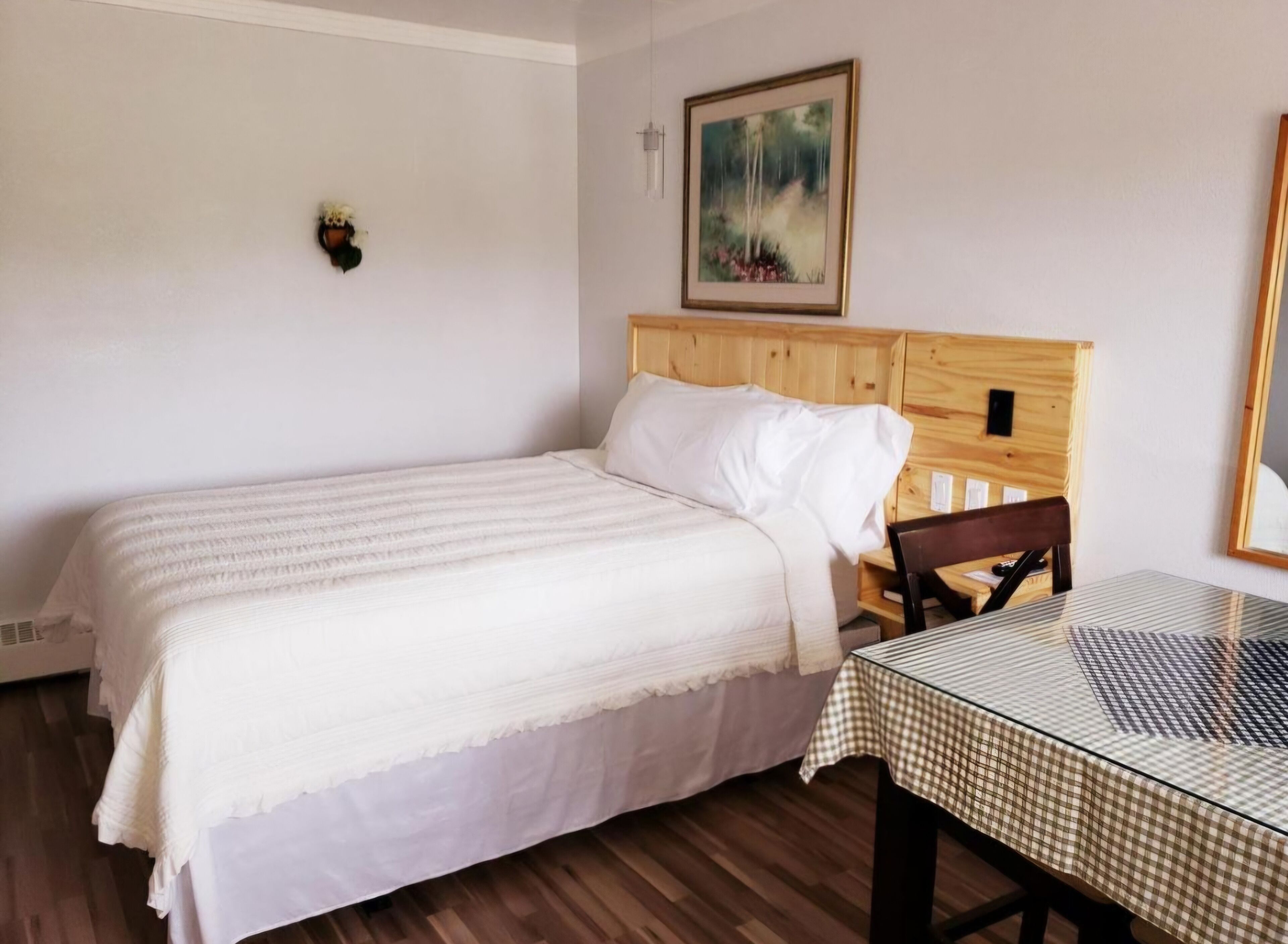 Une chambre à coucher avec un lit, une tête de lit en bois, une table recouverte d’une nappe à carreaux, une lampe fixée au mur et un tableau encadré.