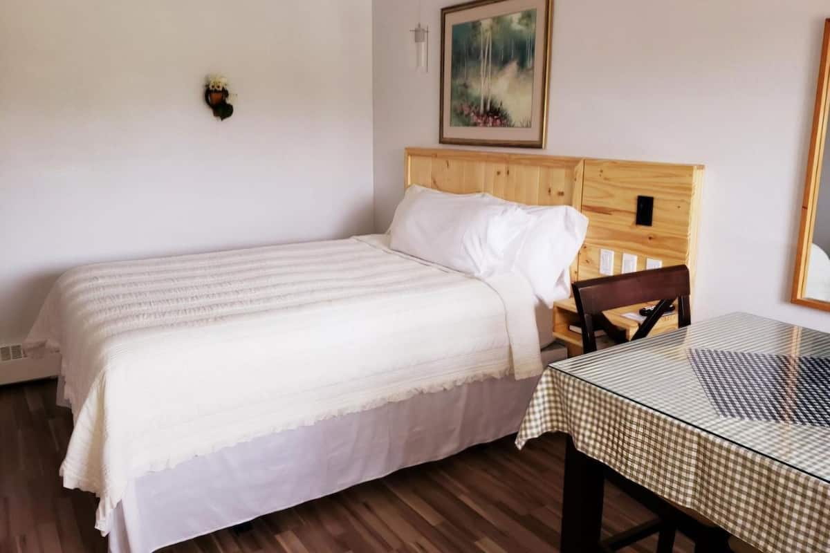 Quarto com cama, cabeceira de madeira, mesa com toalha xadrez, luminária fixada na parede e quadro emoldurado.