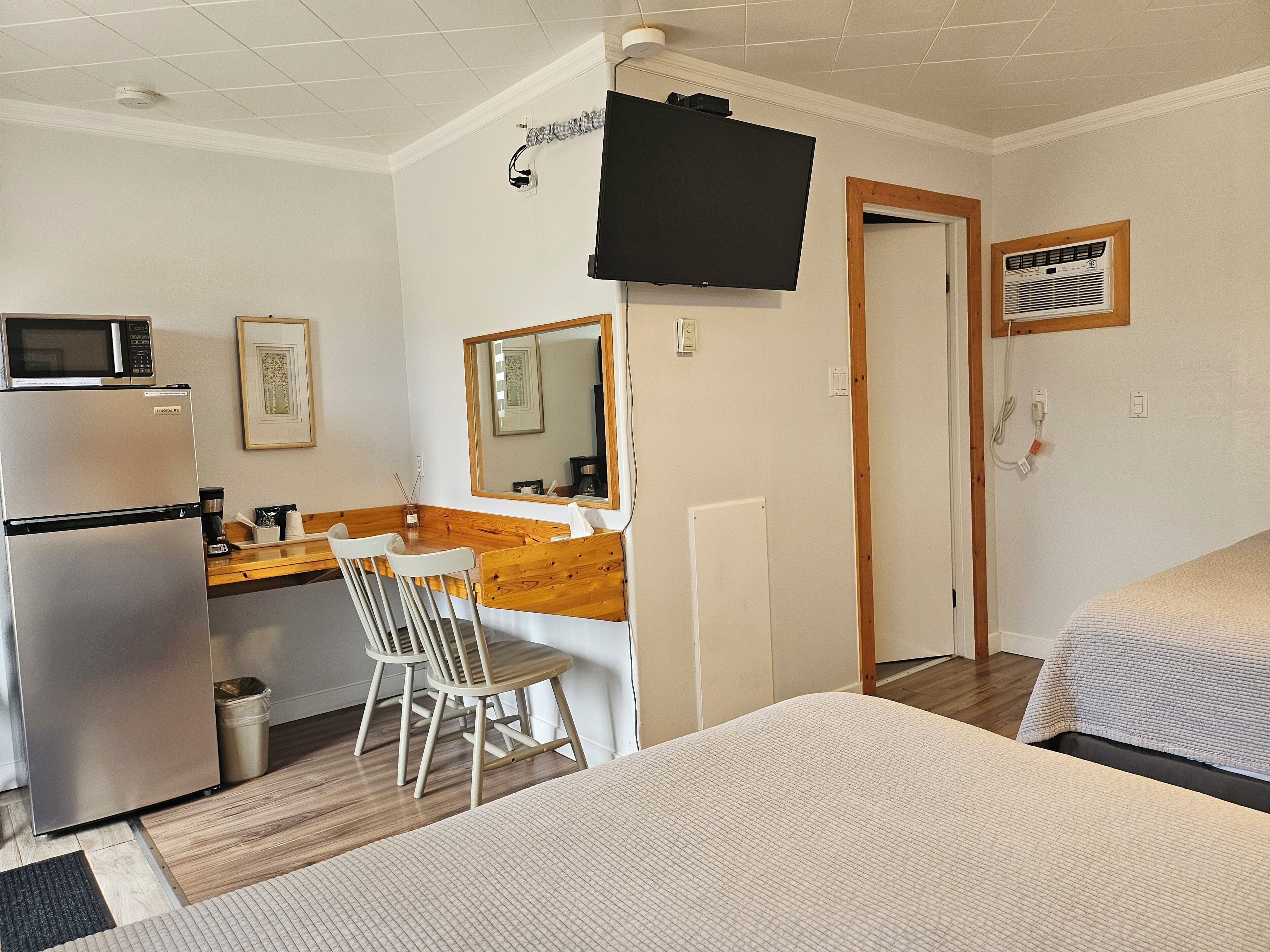 Ein Hotelzimmer mit einem Bett, einem Schreibtisch mit Stuhl, einer Mikrowelle, einem Kühlschrank, einem Fernseher und einem Fenster mit Klimaanlage.