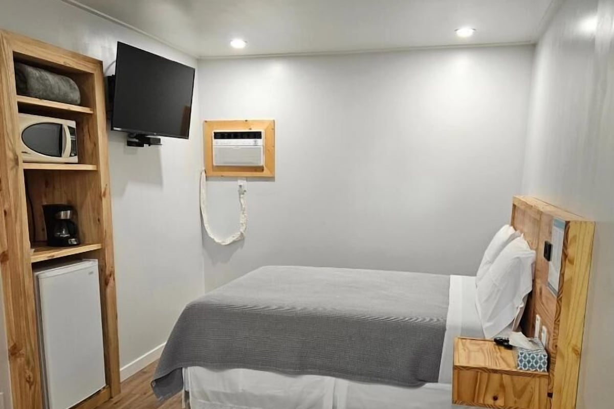 Un dormitorio con cama, televisor, mini refrigerador y estante de madera.