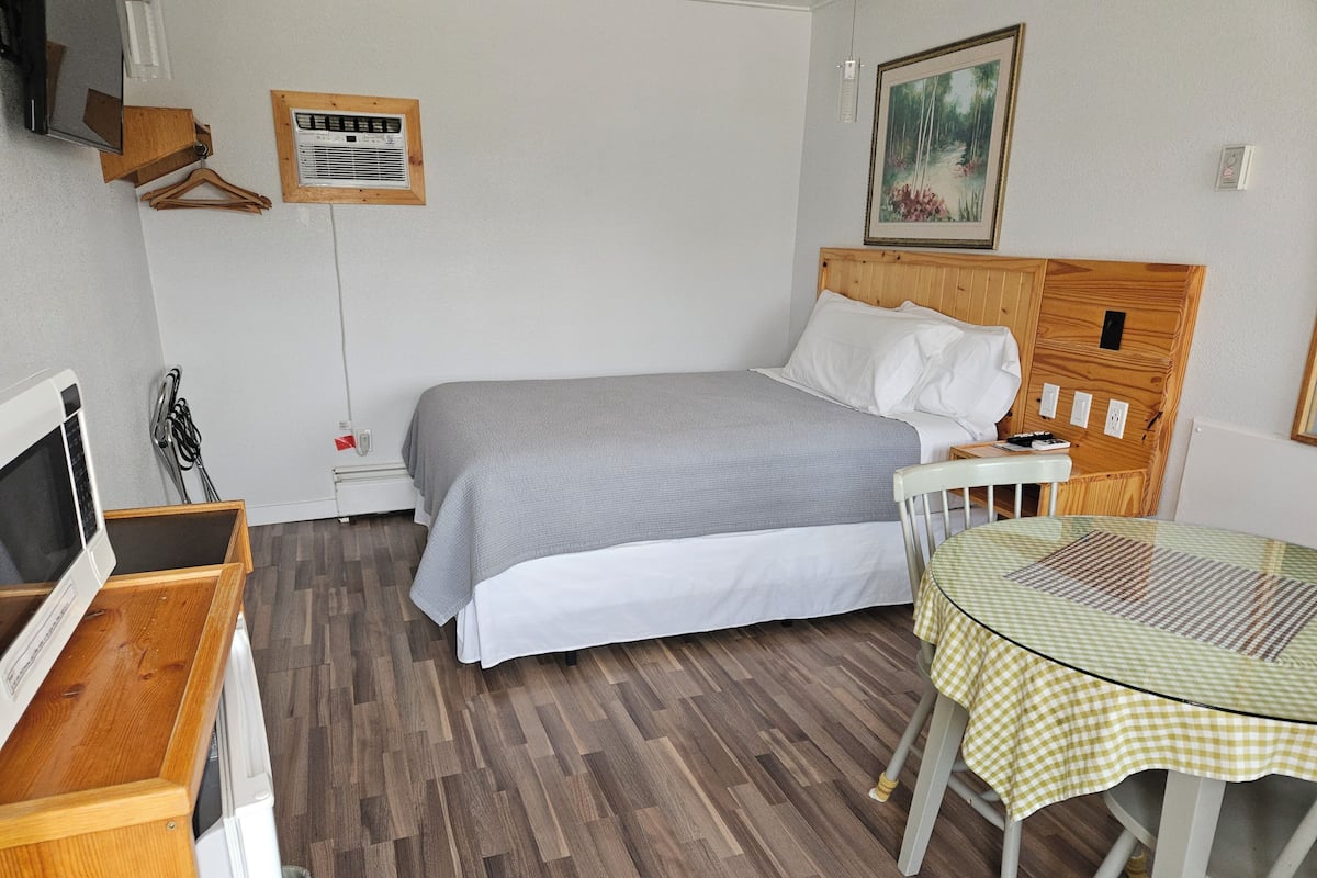 Quarto com cama, mesa de cabeceira, cadeira e uma pequena mesa com toalha xadrez.