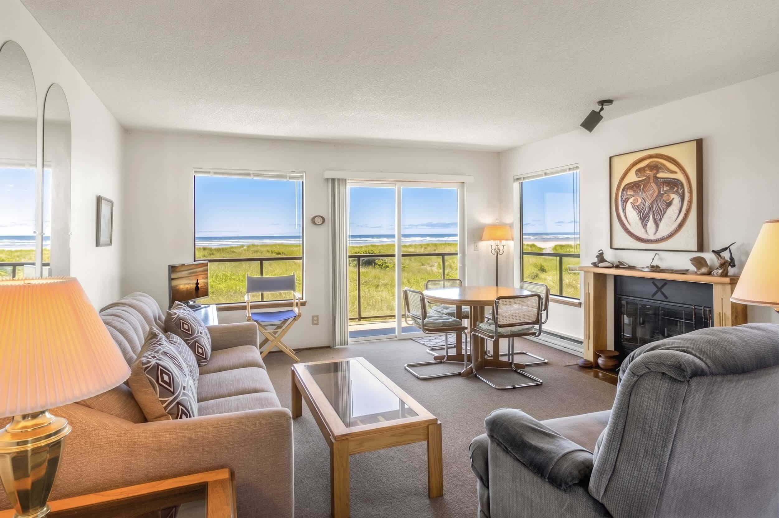 A spacious living room with a sofa, coffee table, and a fireplace. Large windows offer views of the outdoors.