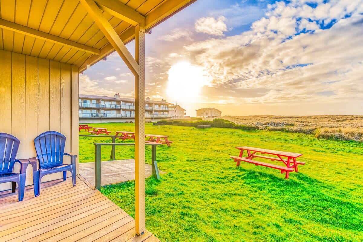 Un espacio al aire libre cubierto con mesas y sillas de picnic, un campo de césped y edificios al fondo.