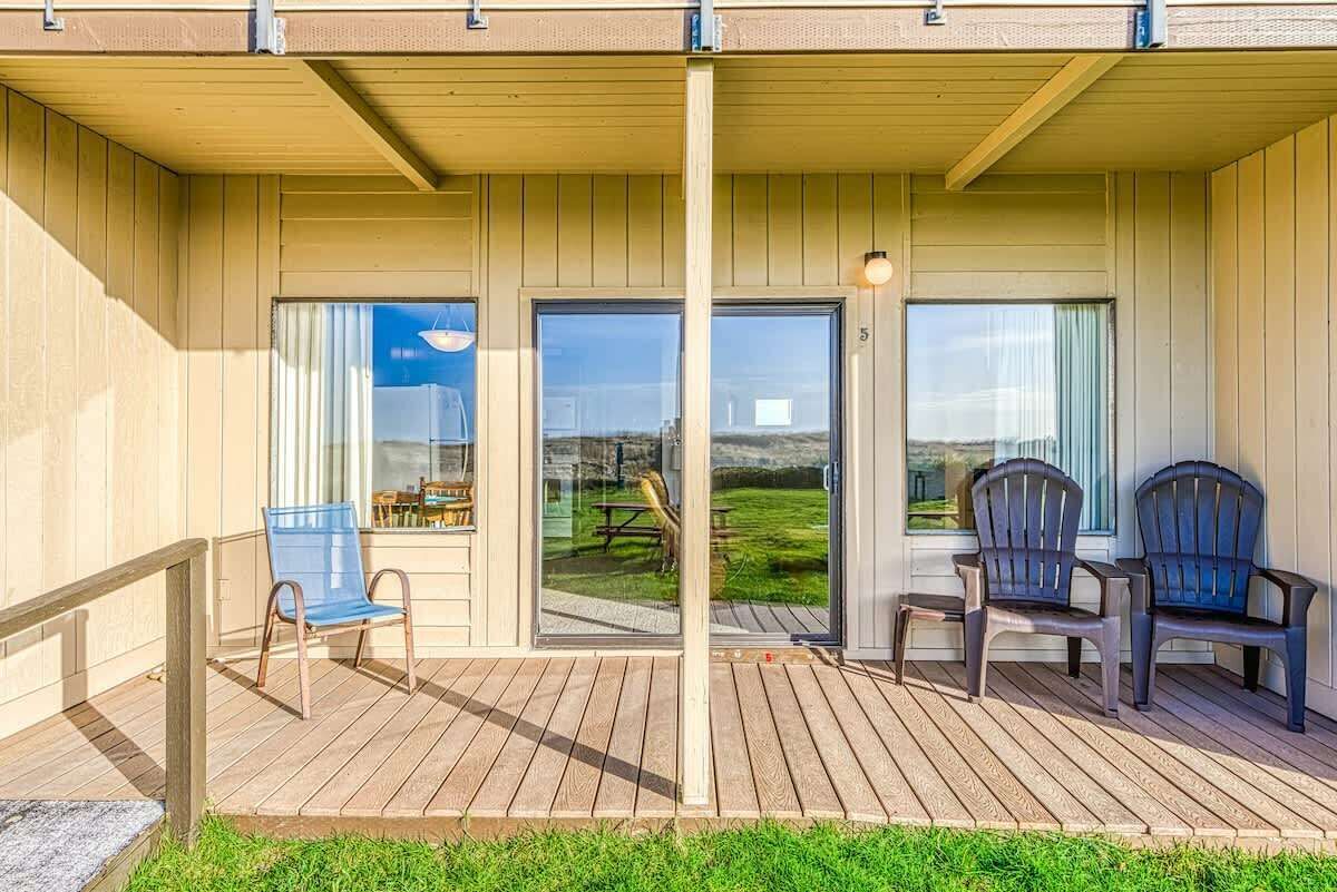 A covered outdoor area with wooden flooring, two chairs, and a view of a grassy field through glass doors.