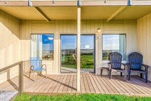 A covered outdoor area with wooden flooring, two chairs, and a view of a grassy field through glass doors.