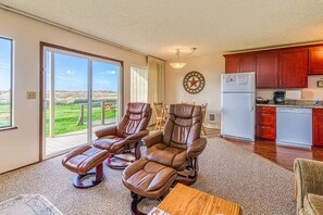 A living room with leather recliners, a dining table, a kitchen with wooden cabinets, and a large sliding glass door leading to a grassy outdoor area.