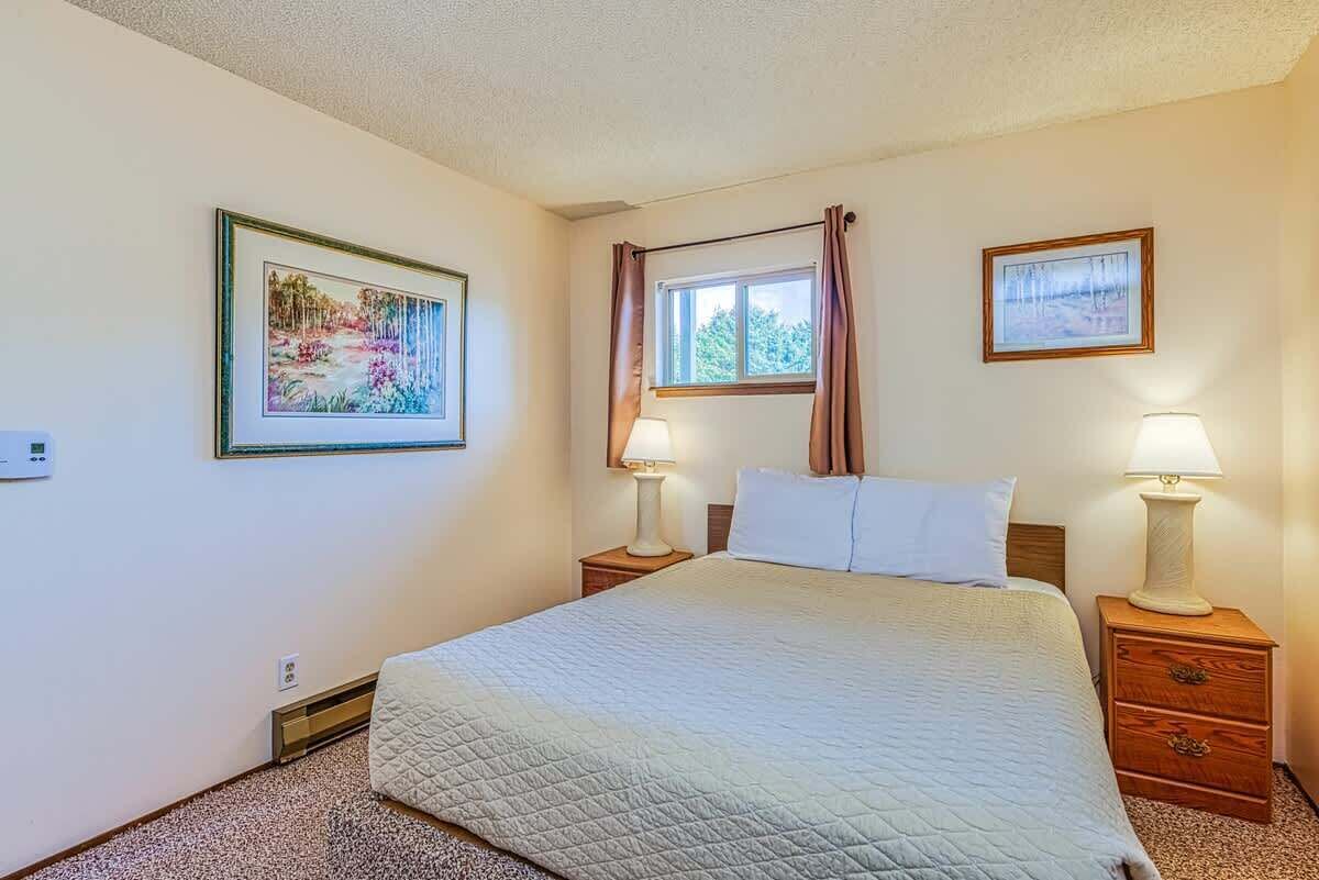 A bedroom with a bed, two bedside tables, two wall-mounted lamps, a window with curtains, and two framed pictures on the walls.