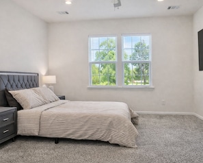 Une chambre à coucher avec un lit, une table de chevet, une lampe et une fenêtre donnant sur des arbres.