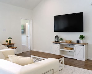 Un salon comprenant un canapé, une table basse, un téléviseur fixé au mur, une petite table d’appoint avec une lampe, et un meuble blanc avec des paniers.