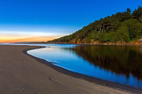 A beach with a calm water body reflecting the sky, a forested hillside, and a clear horizon.
