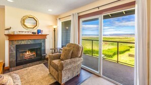 A living room with a fireplace, a sofa, a chair, and a view of a green landscape through large sliding doors. - Hi-Tide Resort, Top Floor, Queen Bed, Unit 29 (Moclips)