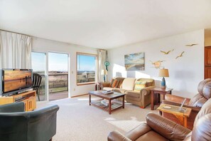 A spacious living room with a sofa set, a coffee table, a TV, and a view through large windows. - Hi-Tide Resort, Queen Bed, Unit 12 (Moclips)
