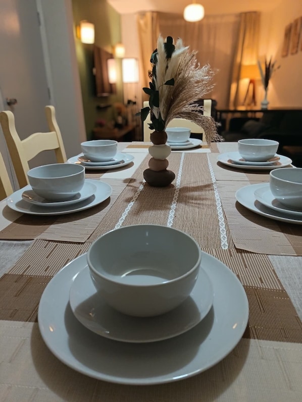 Une table à manger dressée avec des bols et des assiettes blancs, un vase décoratif contenant des plantes séchées et un set de table en fibres tressées.