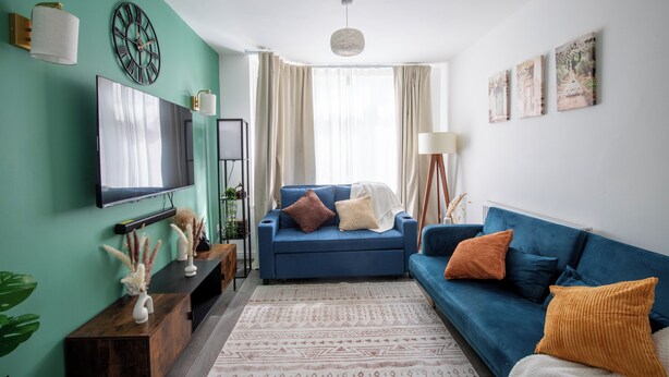 A modern living room with a blue sofa, a flat-screen TV, a large clock, and a window with curtains. - Stylish 3BR Home Near NEC & Airport | Sleeps 6-7 (Solihull)