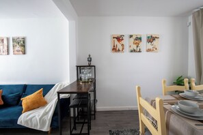 Ein modernes Wohnzimmer mit einem blauen Sofa, einem Esstisch mit Stühlen und einem Couchtisch. An den Wänden hängen drei Bilder mit verschiedenen Motiven.