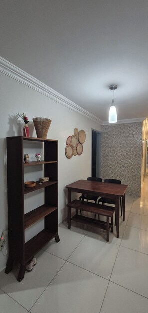 A dining area with a wooden table and benches, a dark wooden bookshelf, and a hanging light fixture. - Comfortable apartment in Maceió (Maceió)