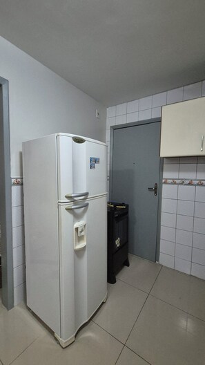 A compact kitchen with a white refrigerator, a small trash bin, and a door leading to another room. - Comfortable apartment in Maceió (Maceió)