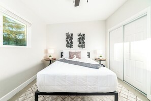 A bedroom with a bed, two bedside tables, a wall art piece, and a view of the outdoors.