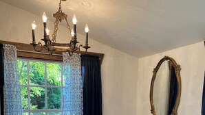 A room with a large window, a round glass table, a bench, a sofa with patterned cushions, a mirror, and a chandelier. - Cozy 1-bedroom cottage in lovely Rocky Mount with AC (Rocky Mount)