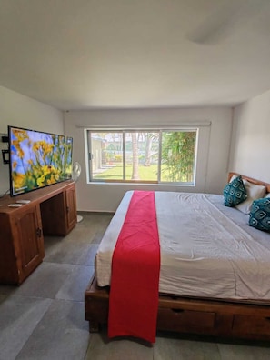 Un dormitorio con cama, un televisor sobre un soporte de madera, una ventana con vistas a la vegetación y una colcha roja.