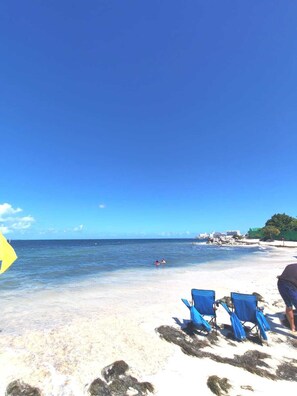 Una playa con cielos despejados, dos sillas azules para la playa y una persona en el agua.